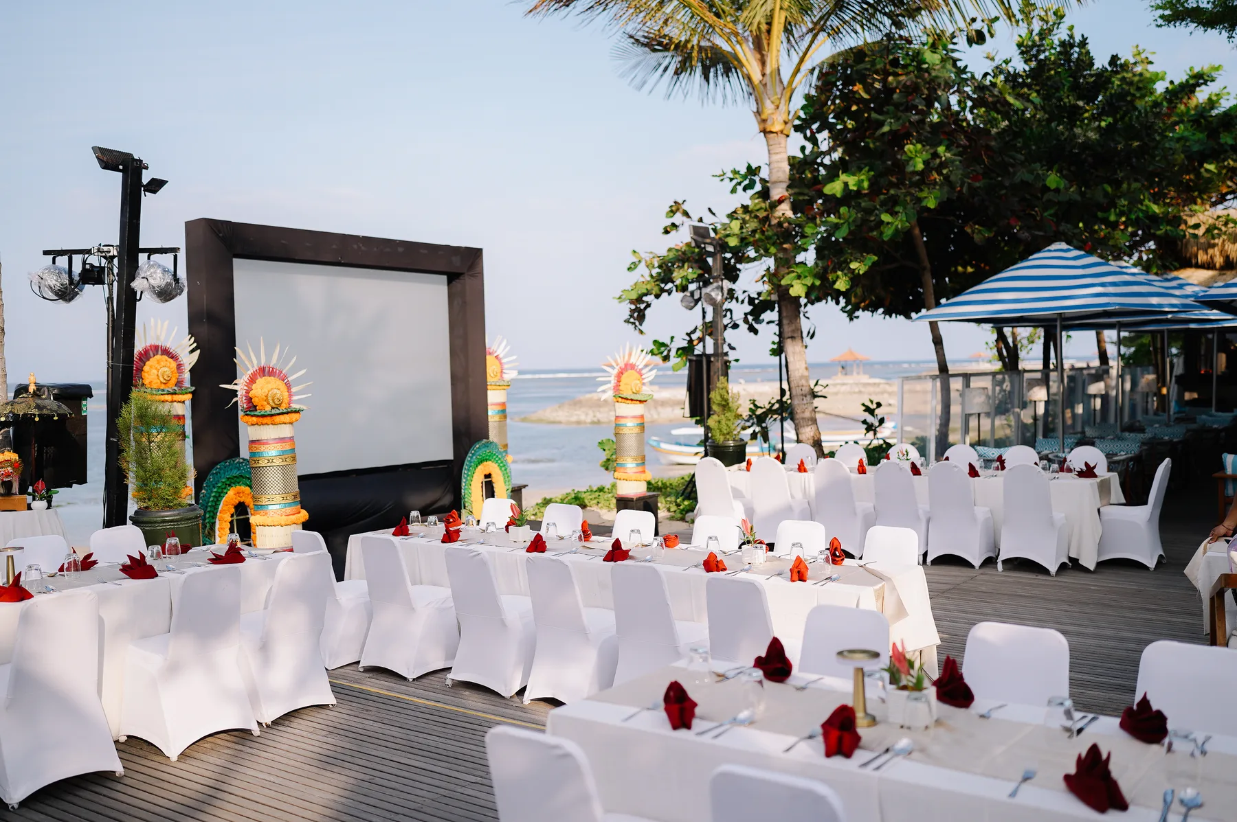 Tables set with white linens and red napkins on a beachfront deck at golden hour, a screen set up nearby
